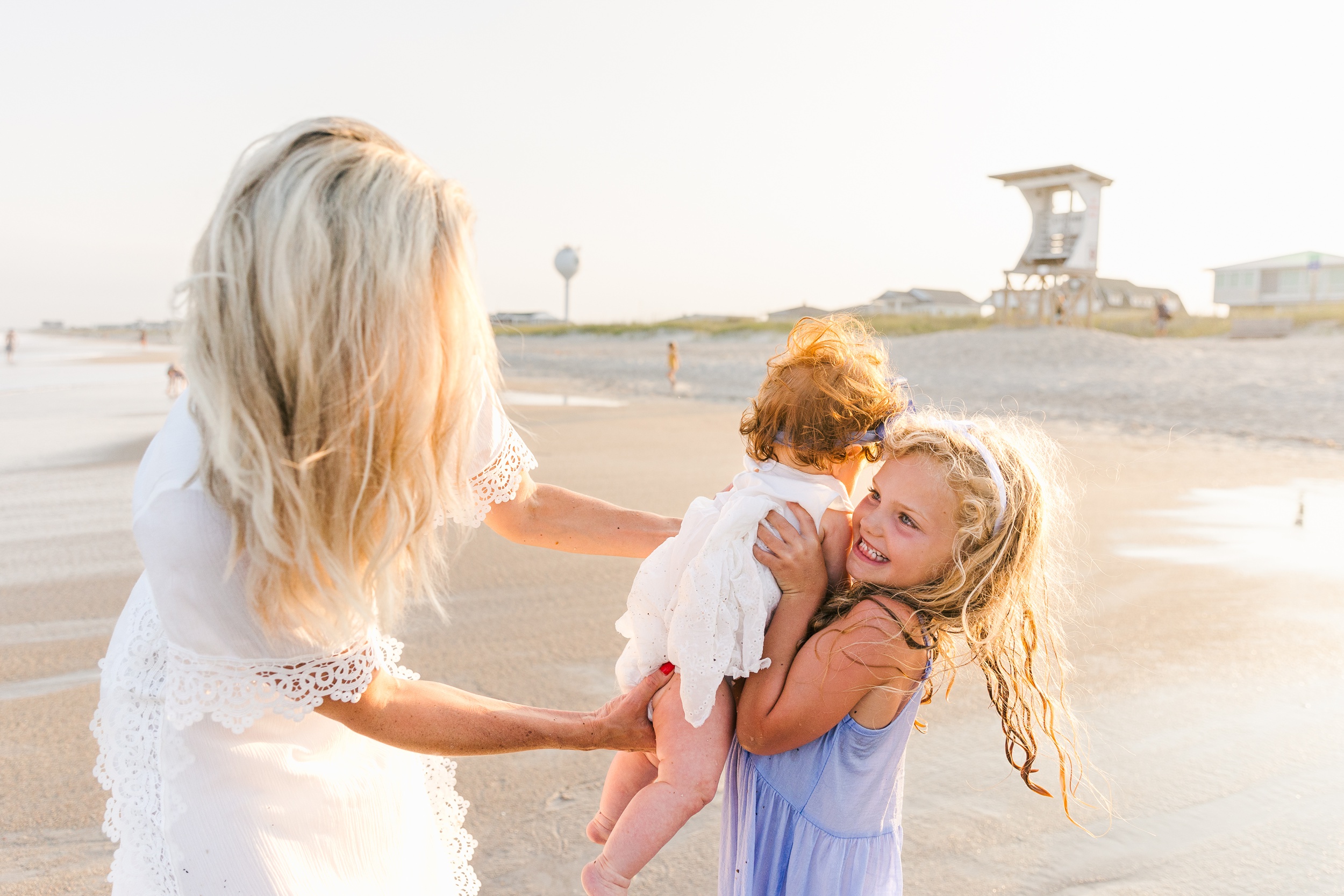 A toddler girl lifts her baby sister while playing on a beach with mom