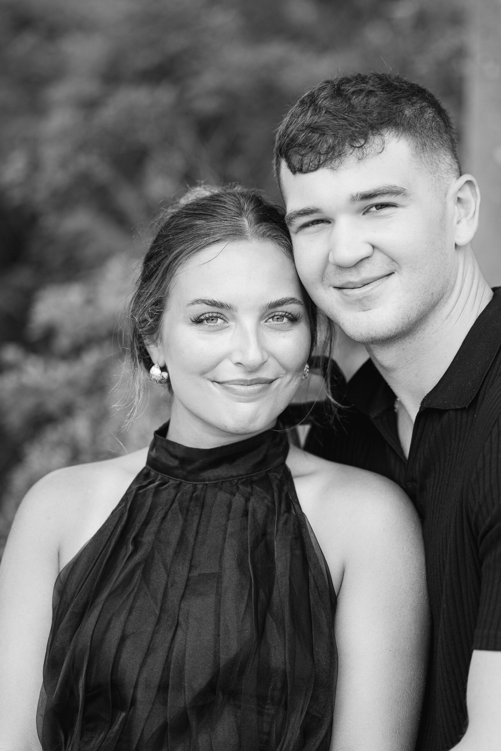 A couple smiles while standing cheek to cheek while wearing black during engagement photos Wilmington NC