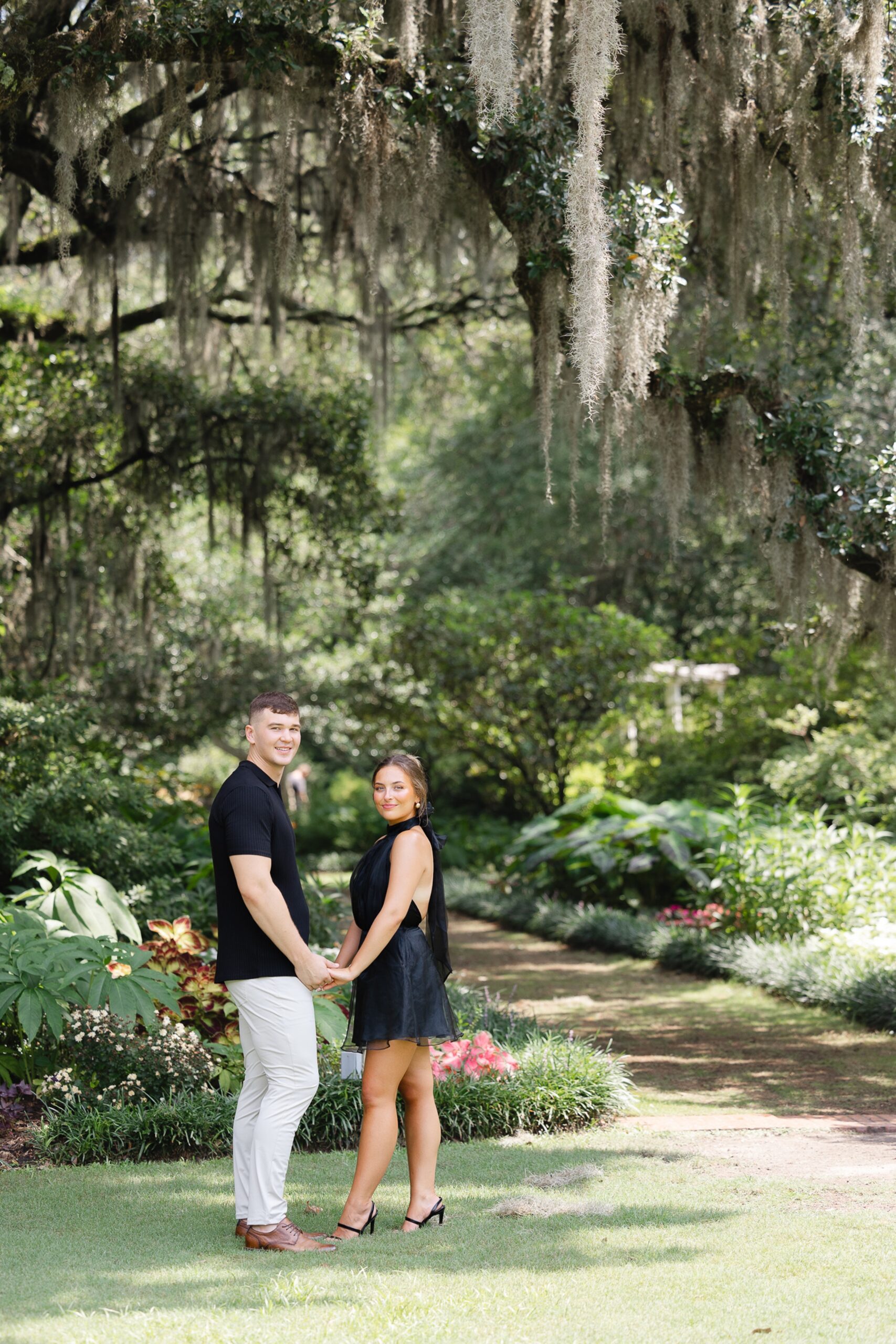 A newly engaged couple stands in a garden lawn holding hands in black