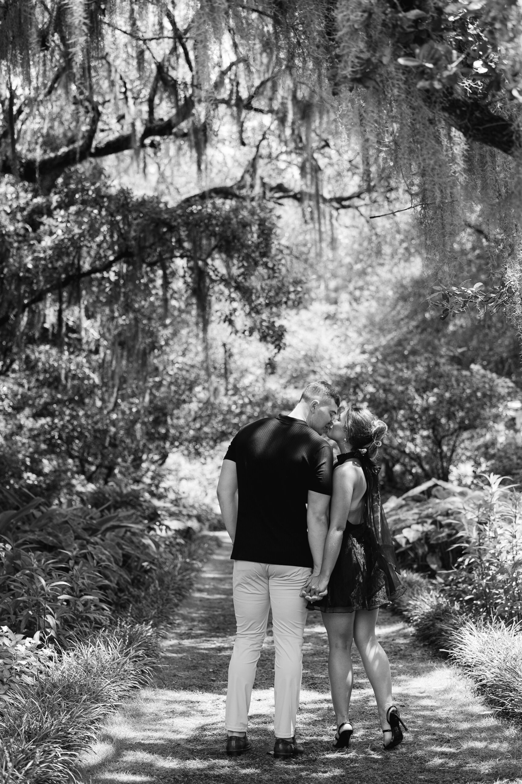 A newly engaged couple in black kiss while walking down a garden trail during their engagement photos Wilmington NC