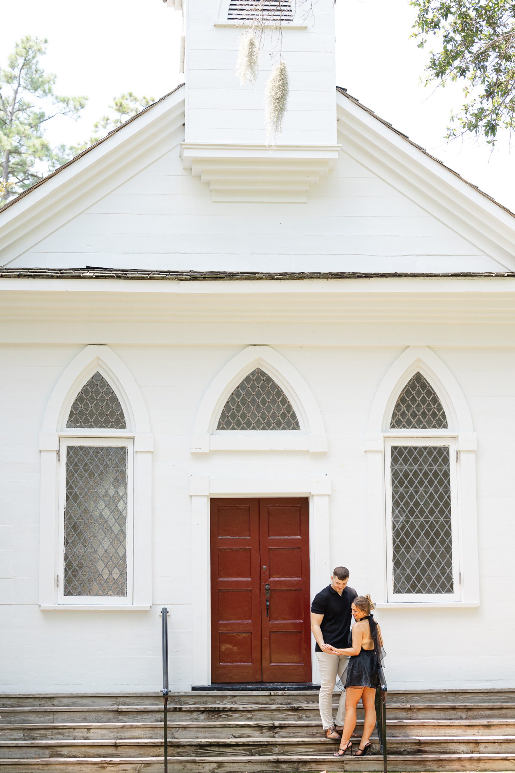 A couple hold hands and admire the engagement ring while standing on steps to an old chapel