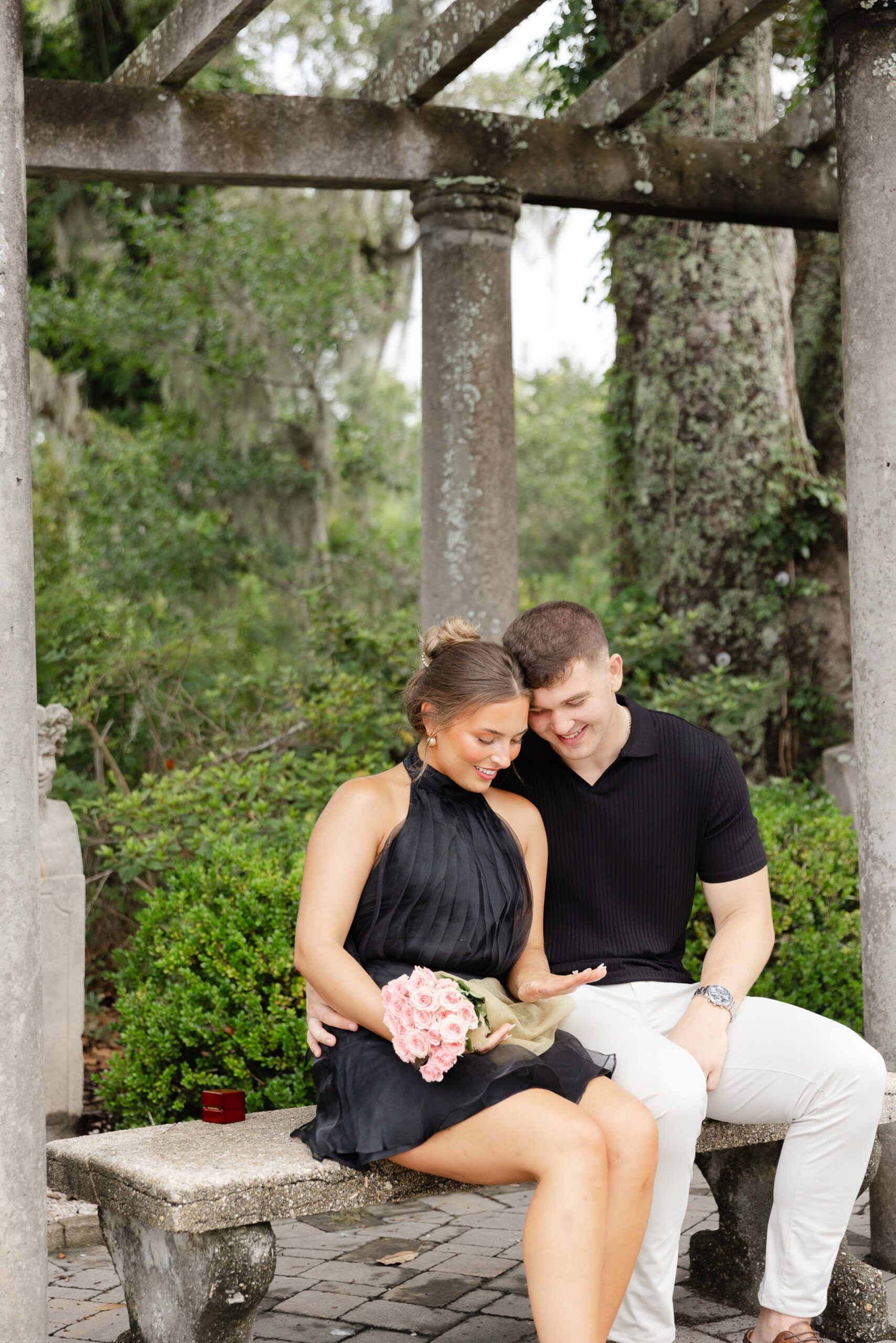 A newly engaged couple sits on a stone garden bench admiring the ring during their engagement photos Wilmington NC