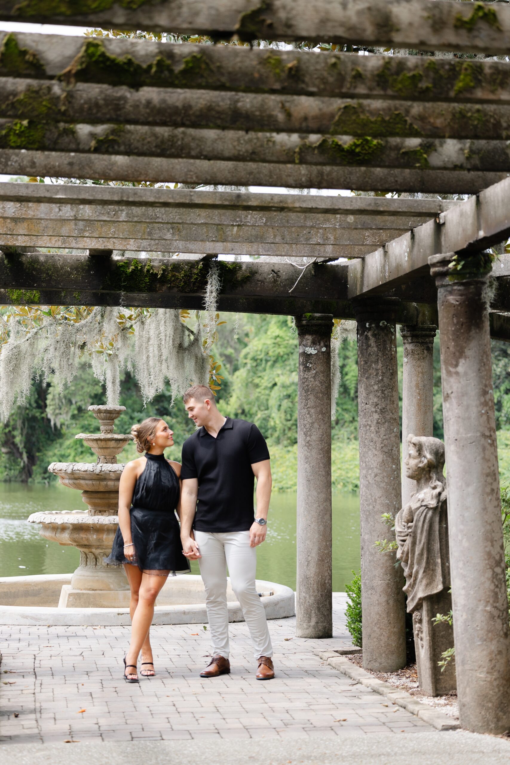 A couple holds hands and walks under a stone pergola with a fountain while wearing black