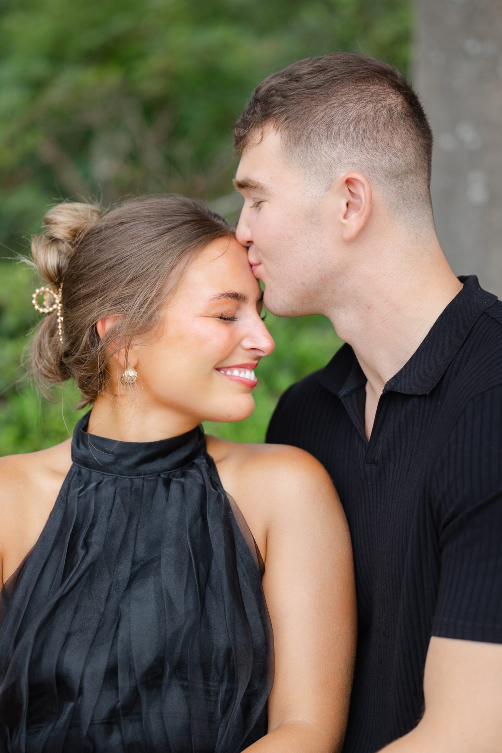 A man kisses his fiancee on the forehead while wearing a black shirt and dress in a garden during engagement photos Wilmington NC