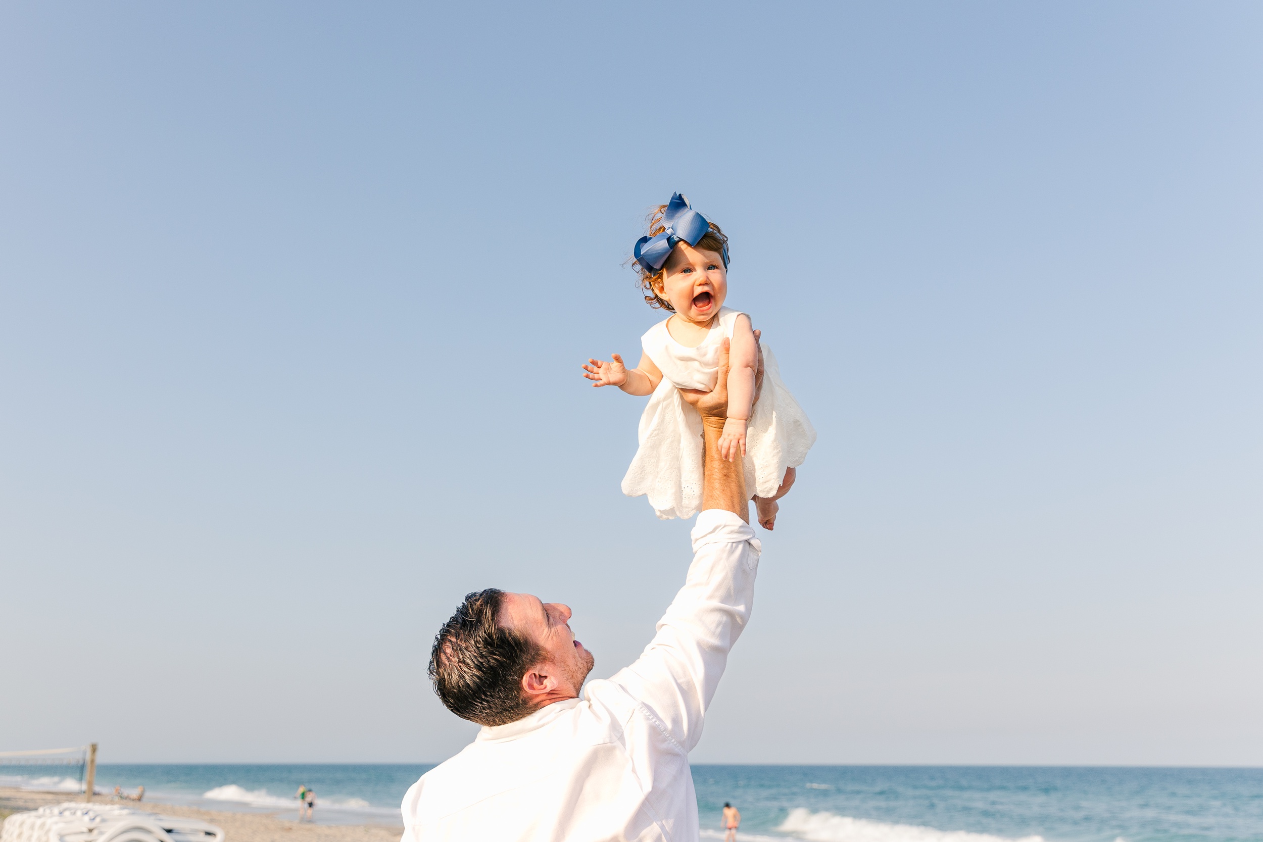 A happy toddler girl in a white dress is lifted into the air by dad with one arm