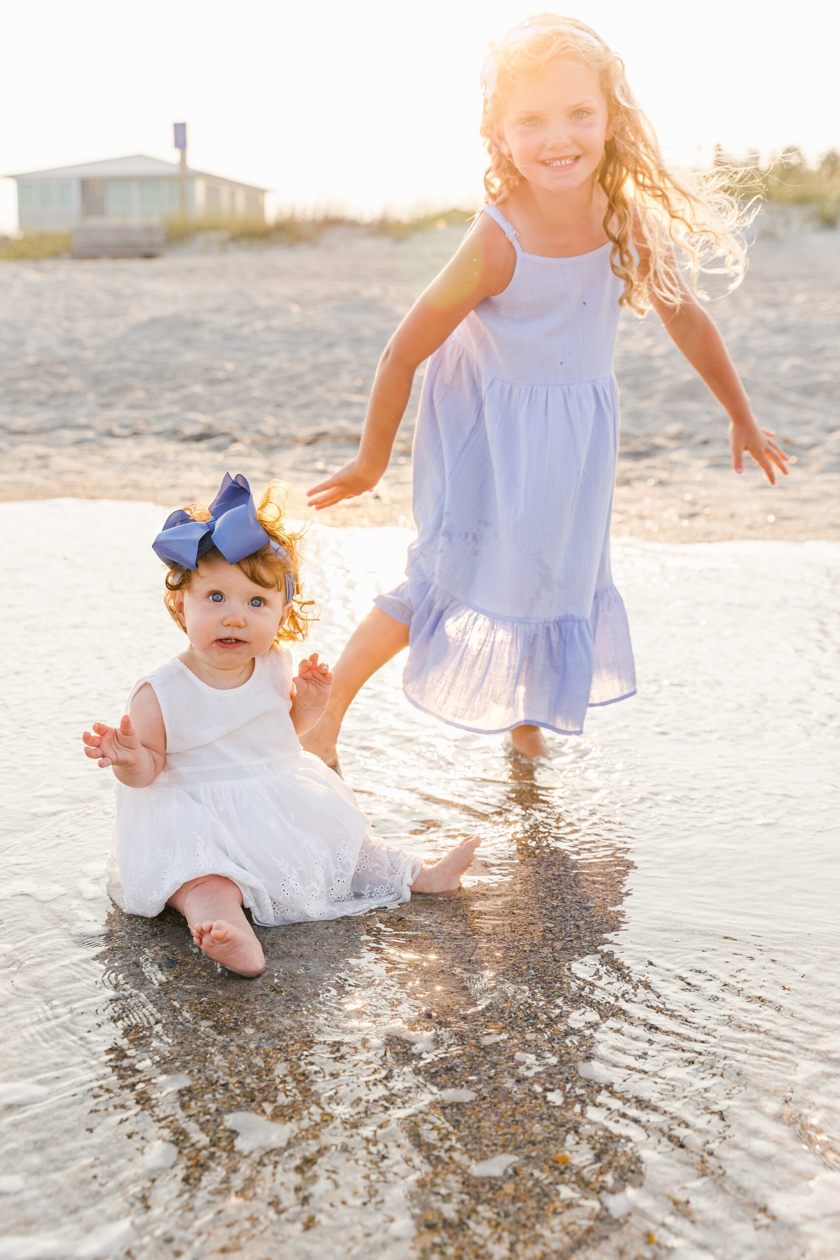 A young girl in a blue dress plays in the water with her baby sister on a beach at sunset