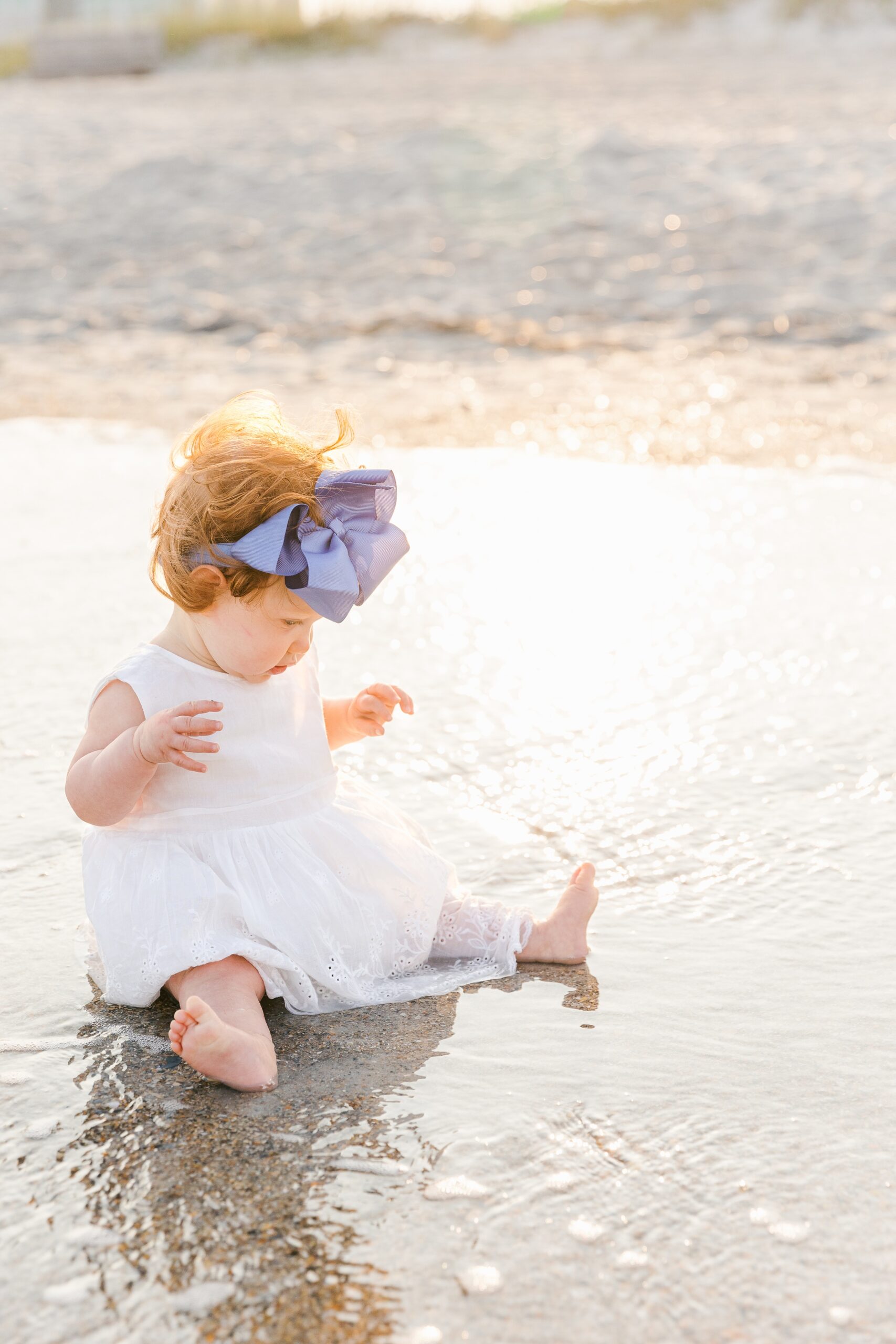 A young toddler girl in a white dress and blue bow explores the water while sitting on a beach at sunset