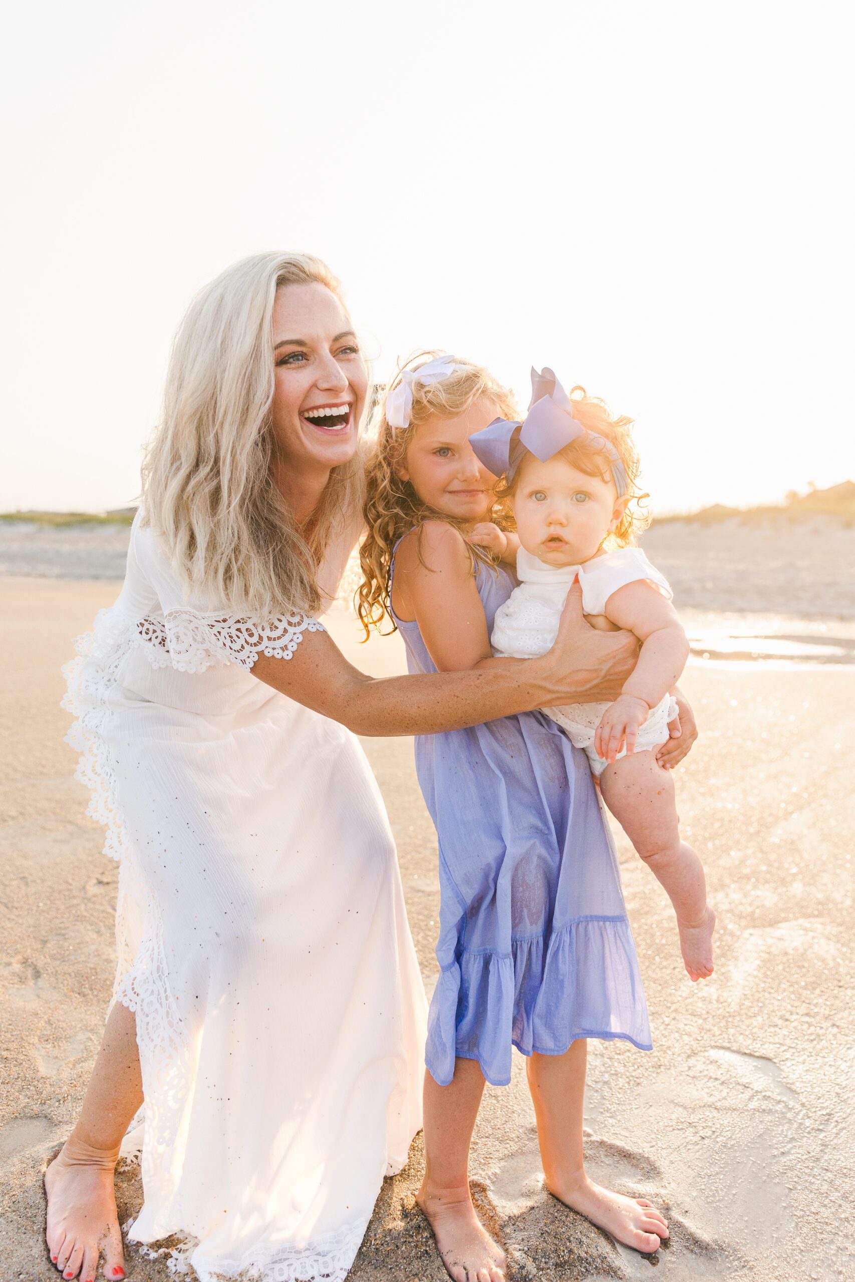 A mom laughs while helping her toddler daughter hold her baby sister on a beach at sunset