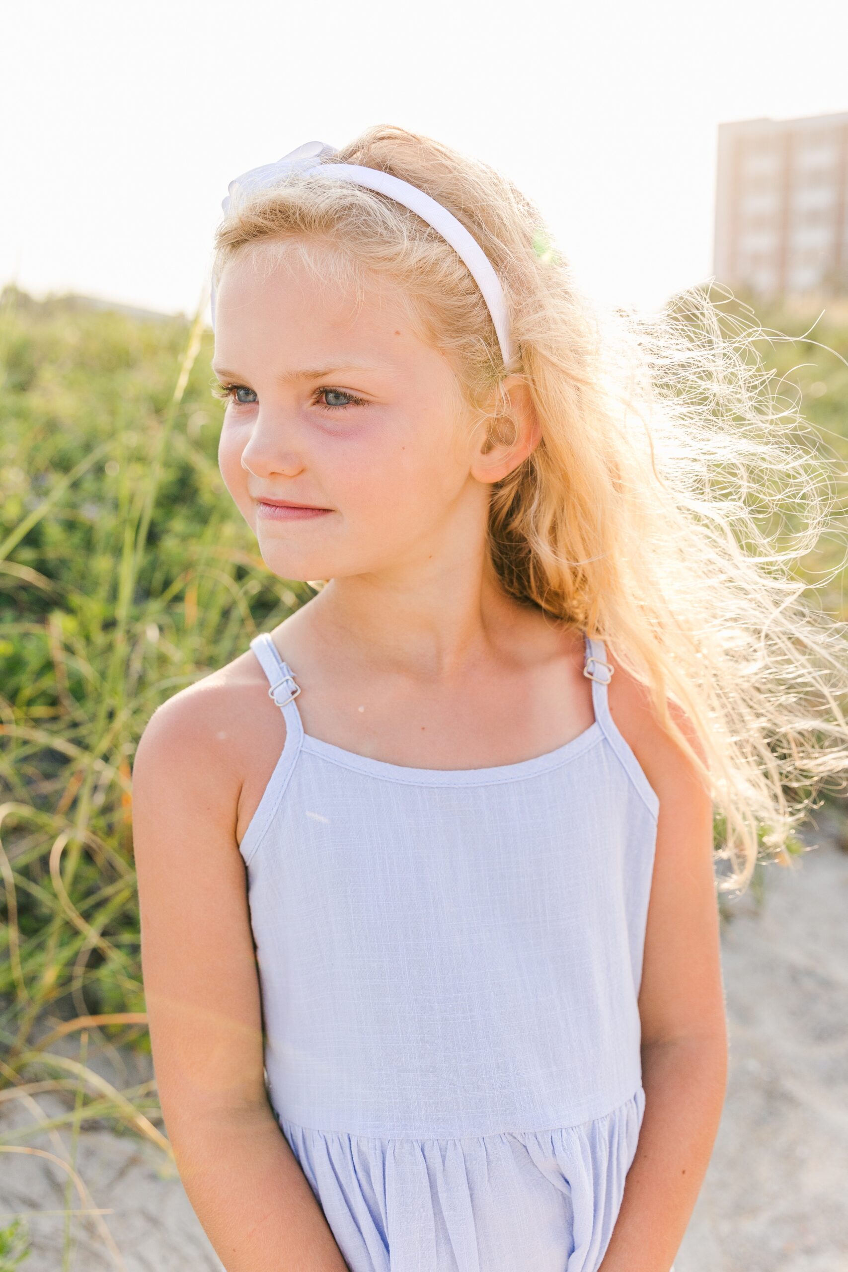 A young girl in a blue dress smiles over her shoulder down the beach while standing on a dune