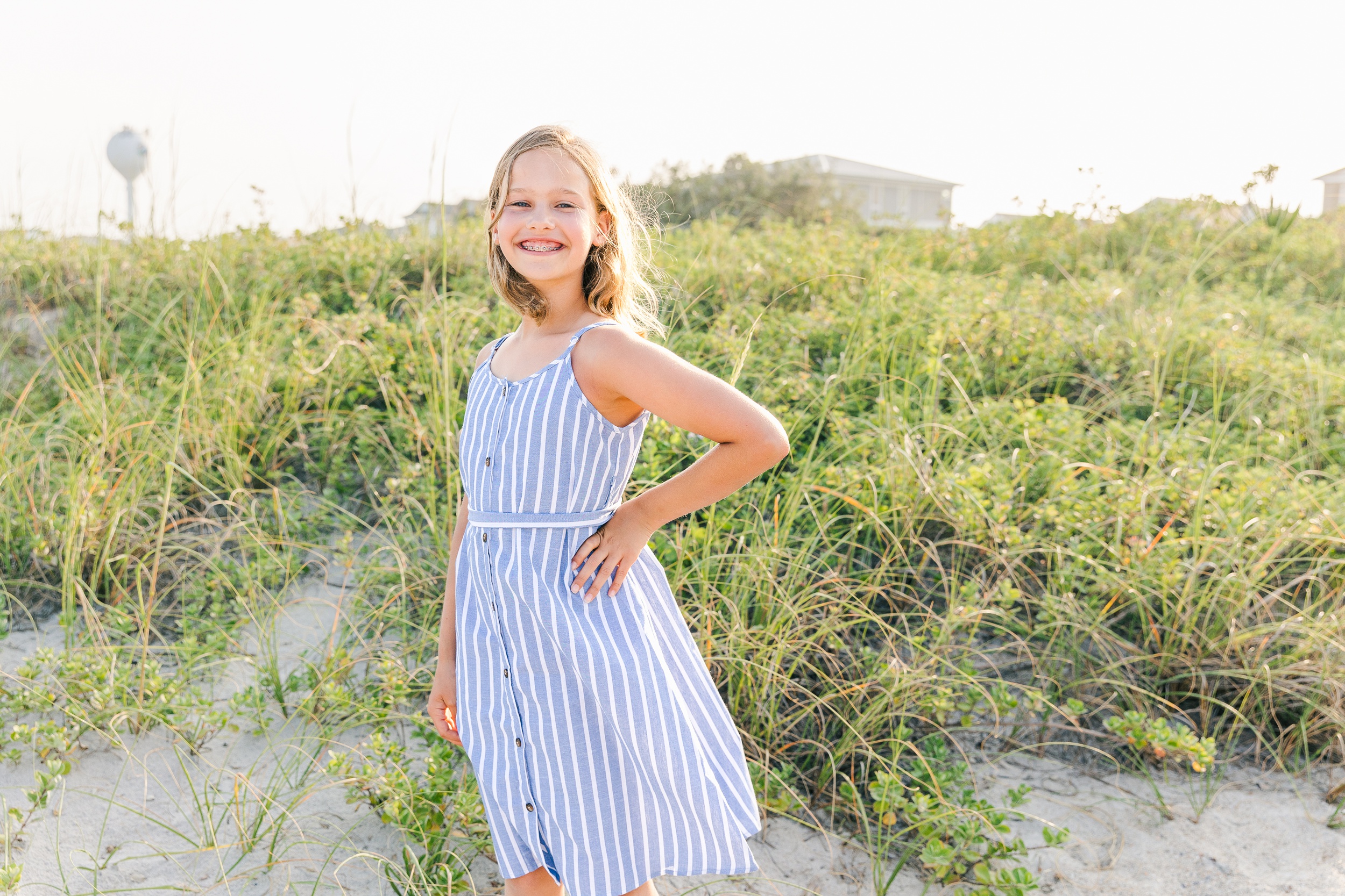 A happy young girl in a blue stripe dress smiles while standing on a windy beach dune