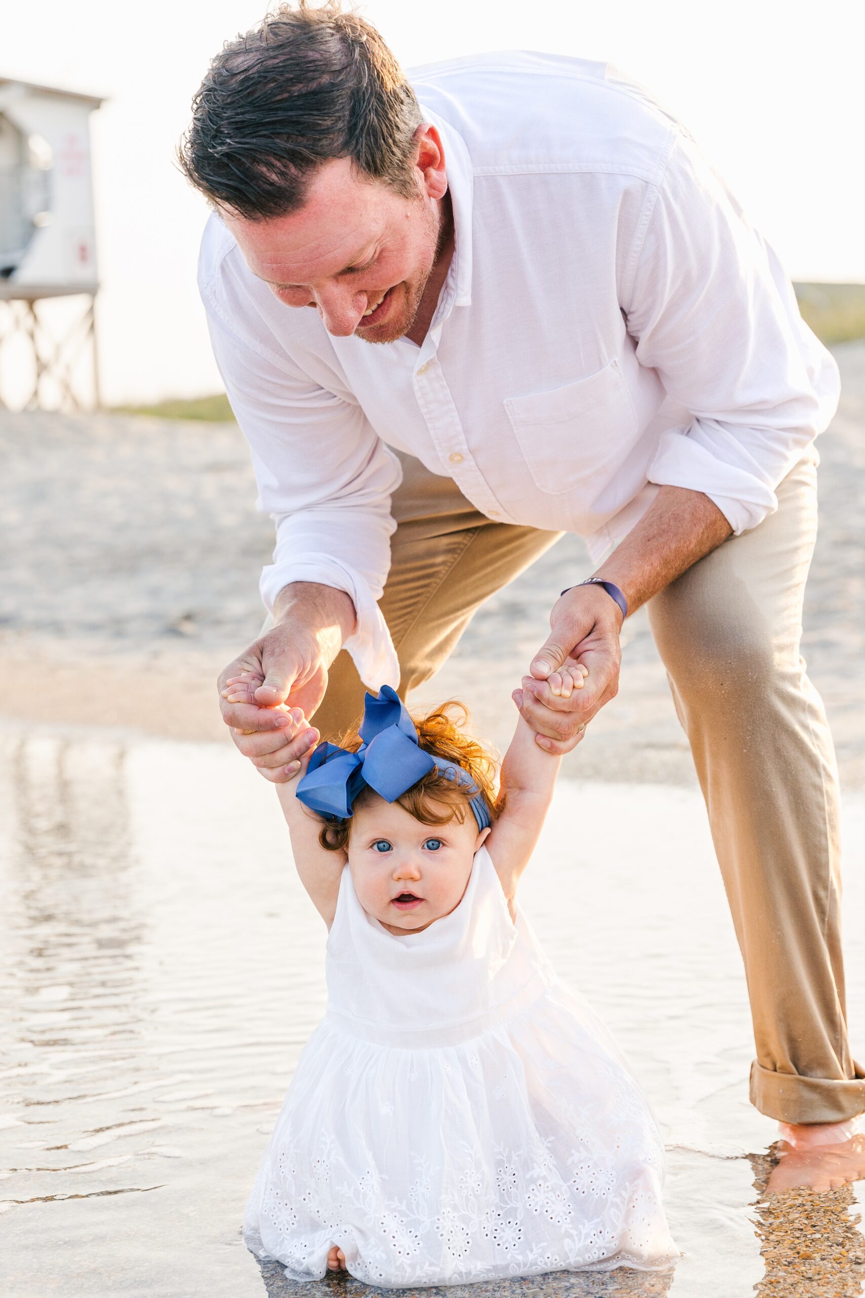 A father in a white shirt plays with his toddler daughter on a beach in the shallow water at sunset during things to do in wilmington with kids