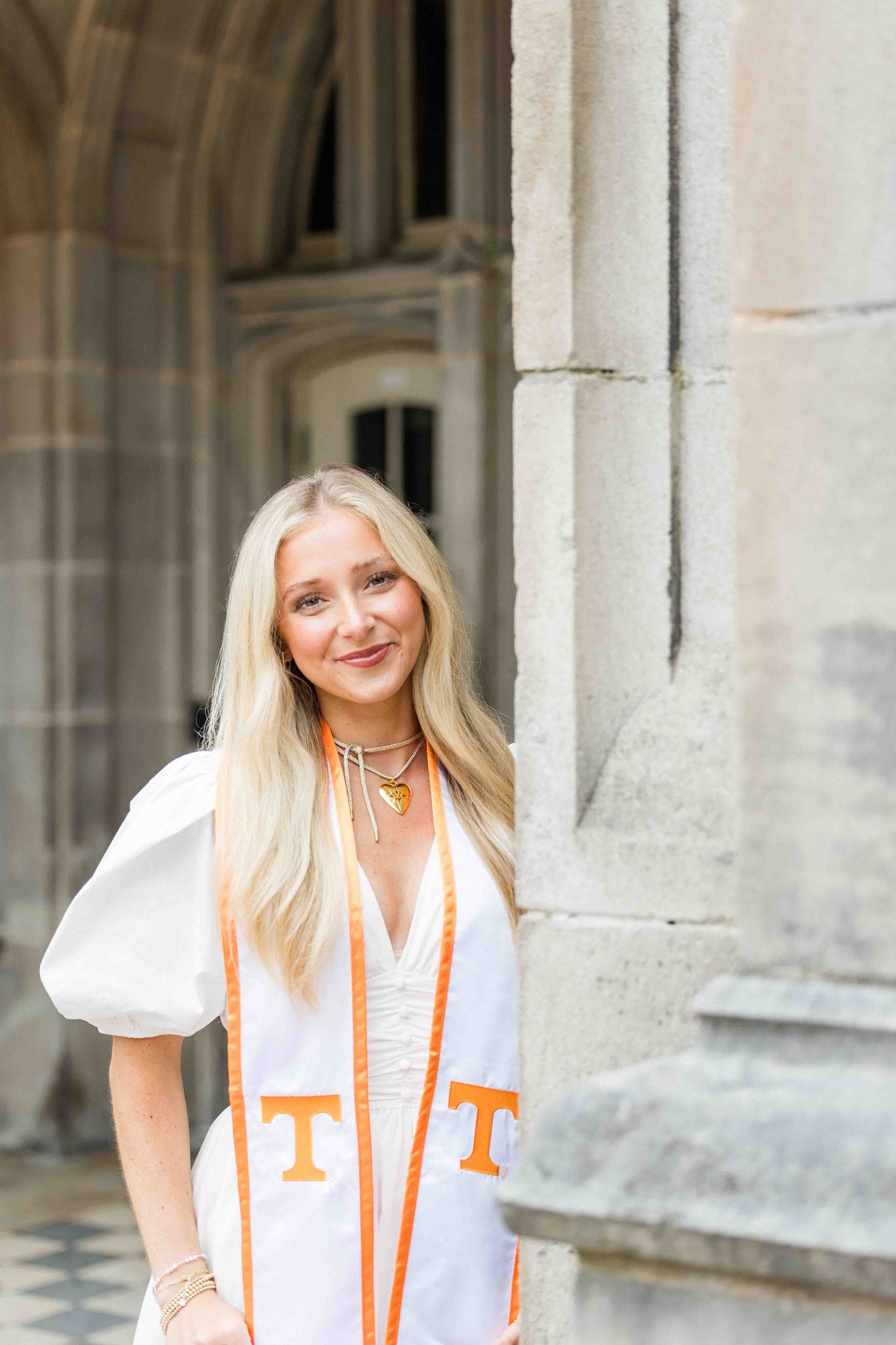 A proud graduate stands at the entrance of an old building smiling in a white dress and stole