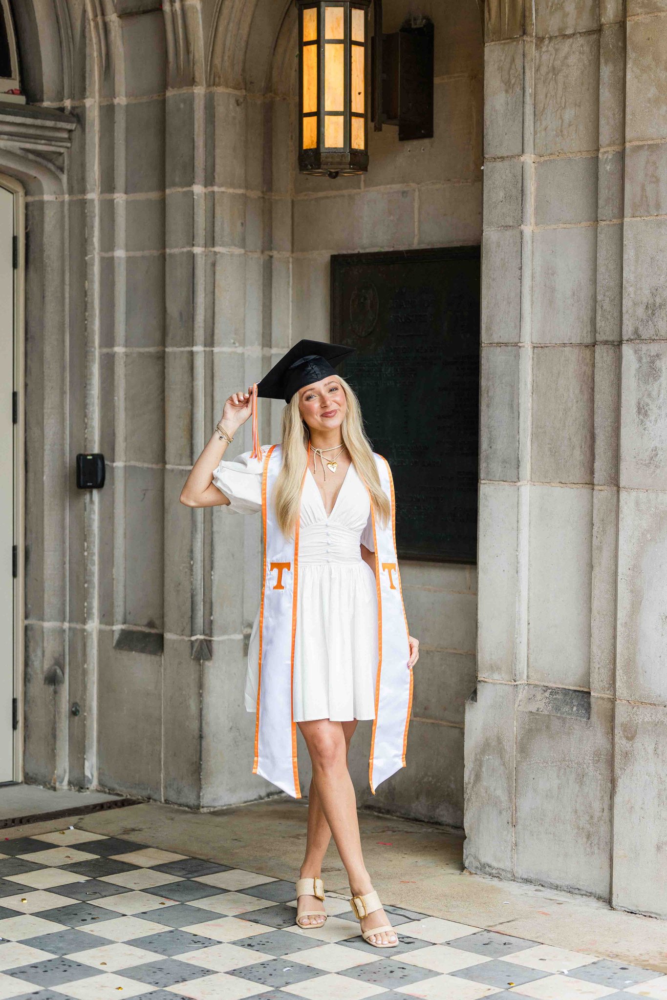 A graduate in a white dress tips her cap while standing in a stone hallway wearing her orange and white stole
