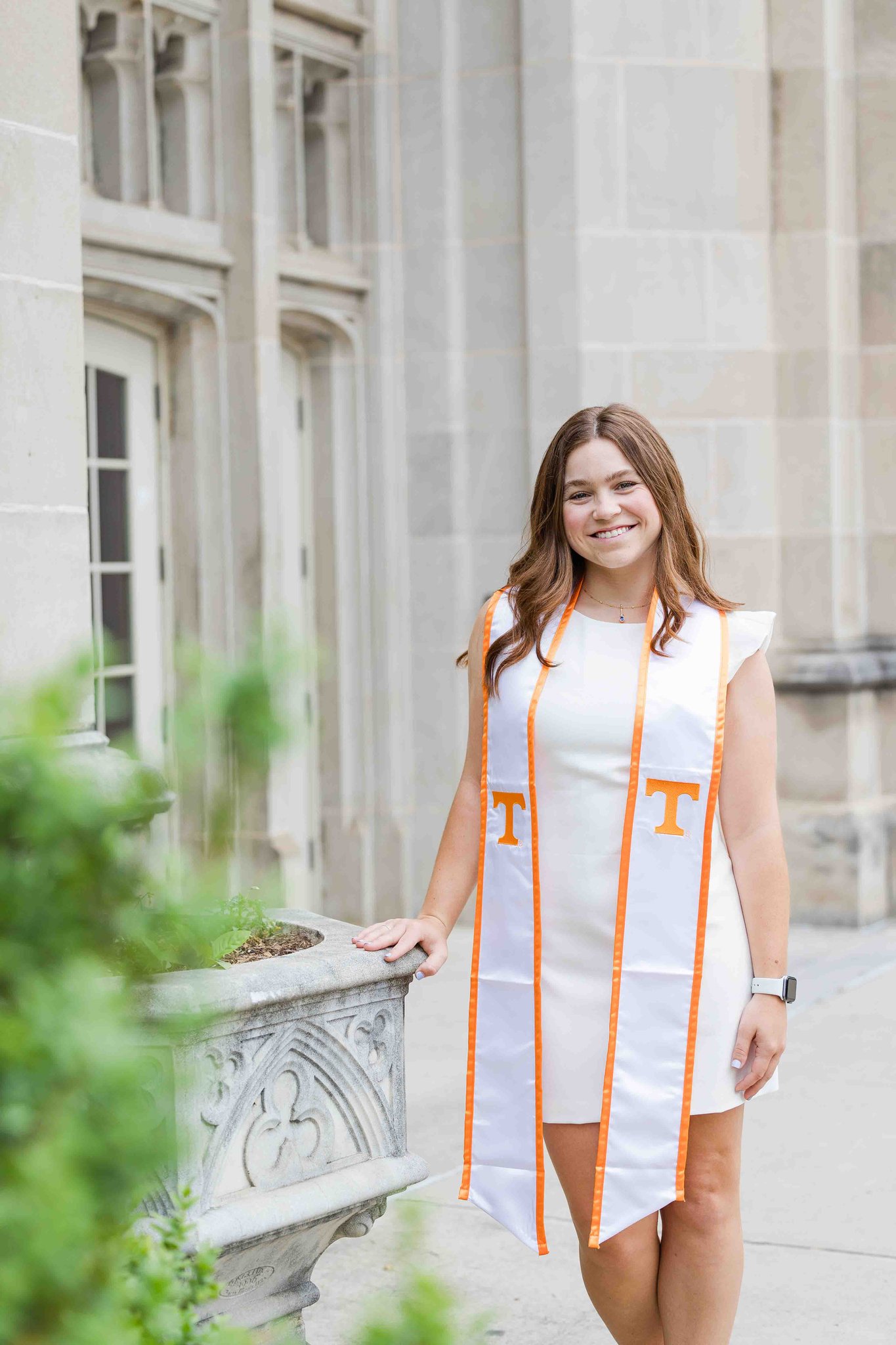 A woman in a white dress leans on a stone planter and smiling in a white dress for her utk graduation pictures