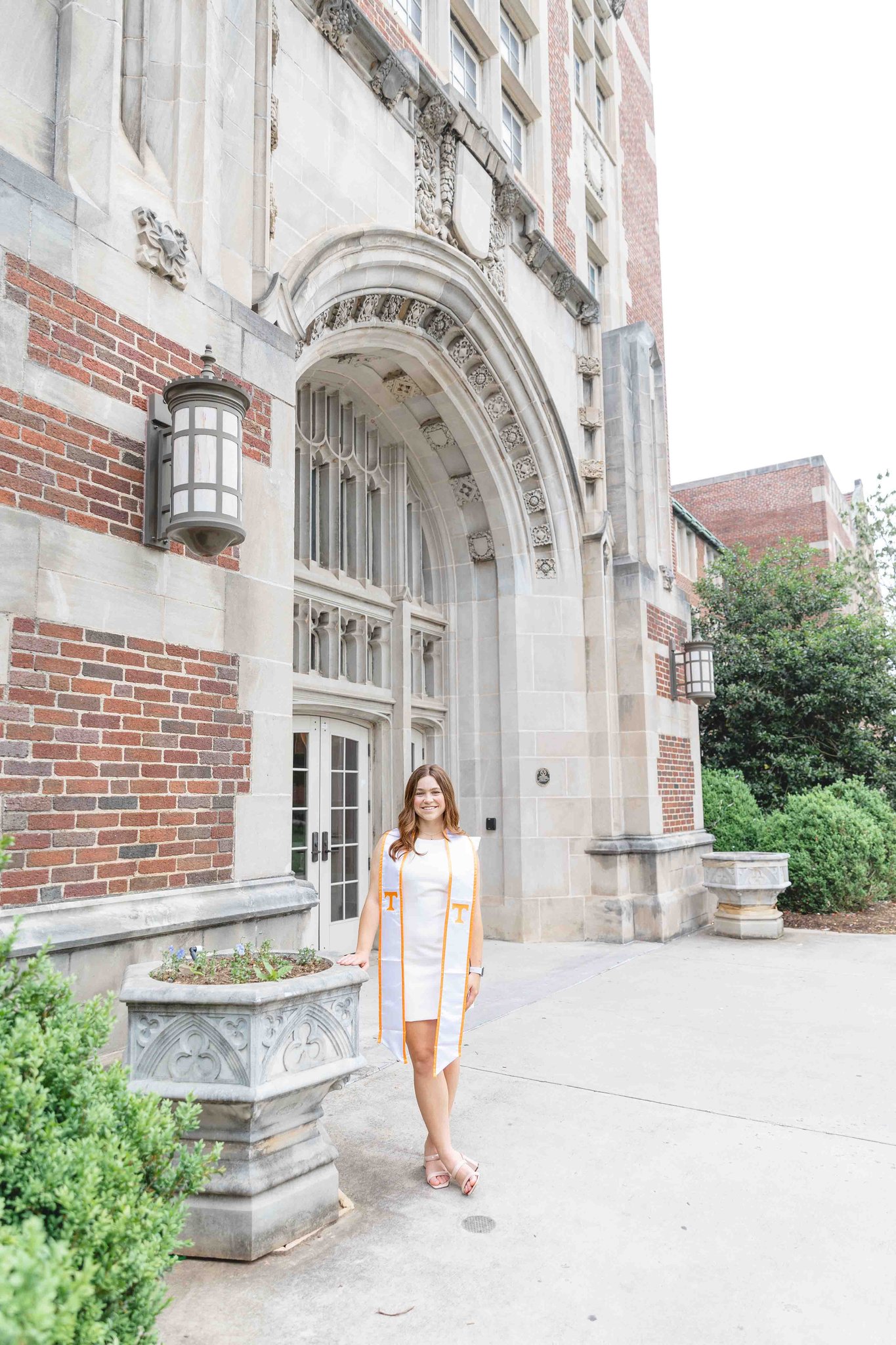 A brunette woman stands against a planter in a white dress and orange stole