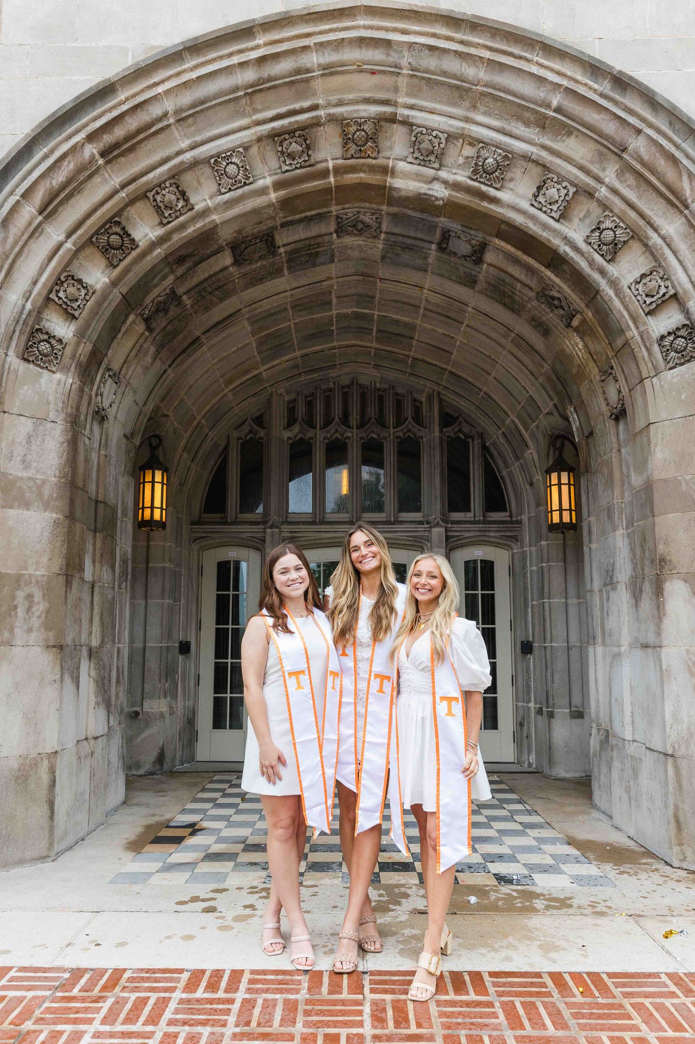 Three women in white dresses and stoles smile together to celebrate their graduation