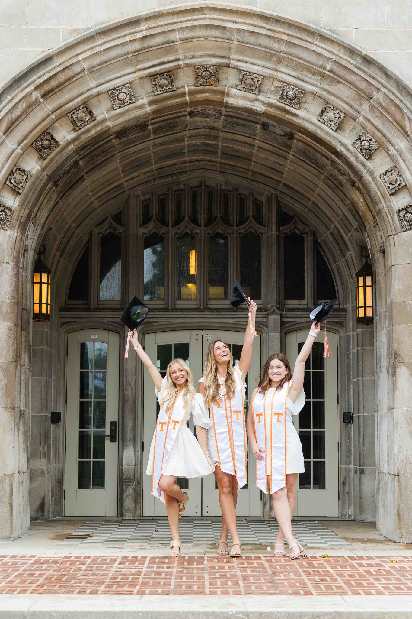 Three woman raise their grad caps in the air while wearing matching white dresses under an ornate arch entrance for their utk graduation pictures