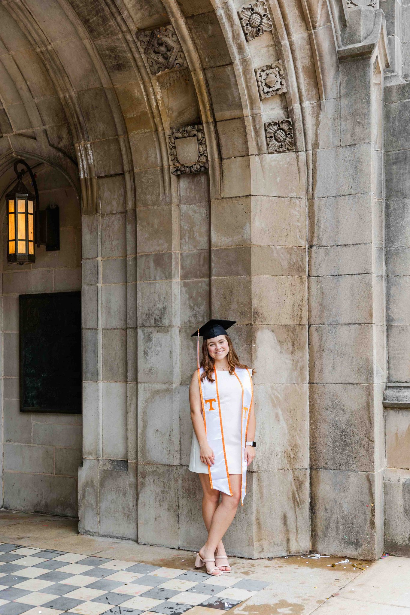 A graduate leans on a wall in a white dress and her cap and stole