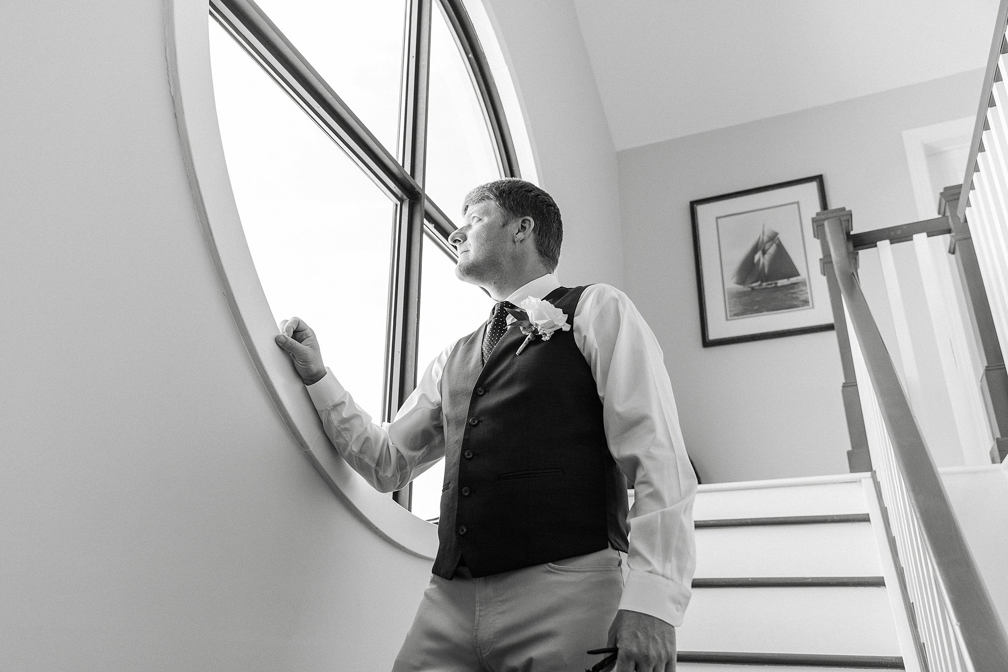 A groom gazes out a round window to the beach after getting ready for his wedding