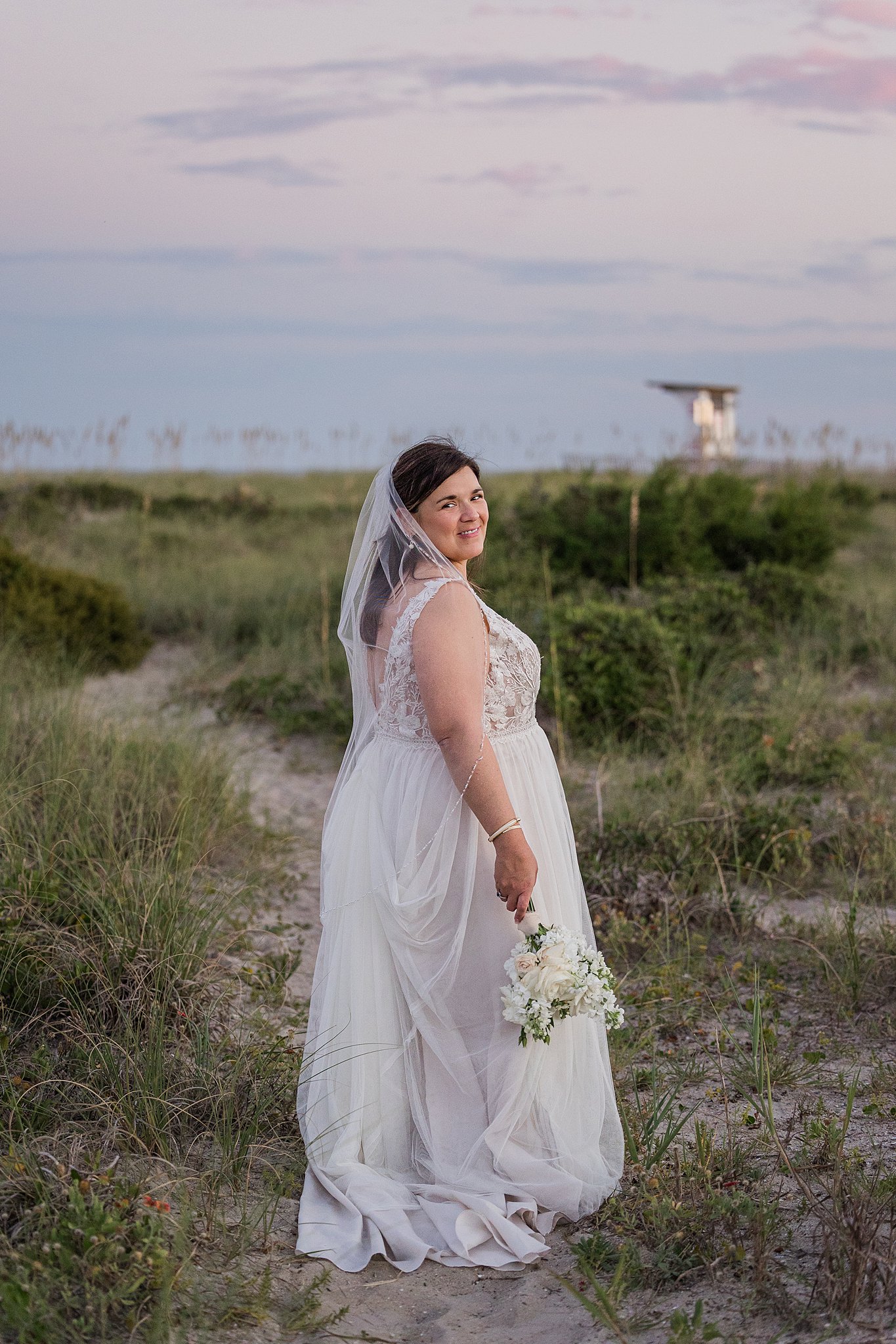 A bride smiles over her shoulder while walking on a beach trail and holding a white rose bouquet