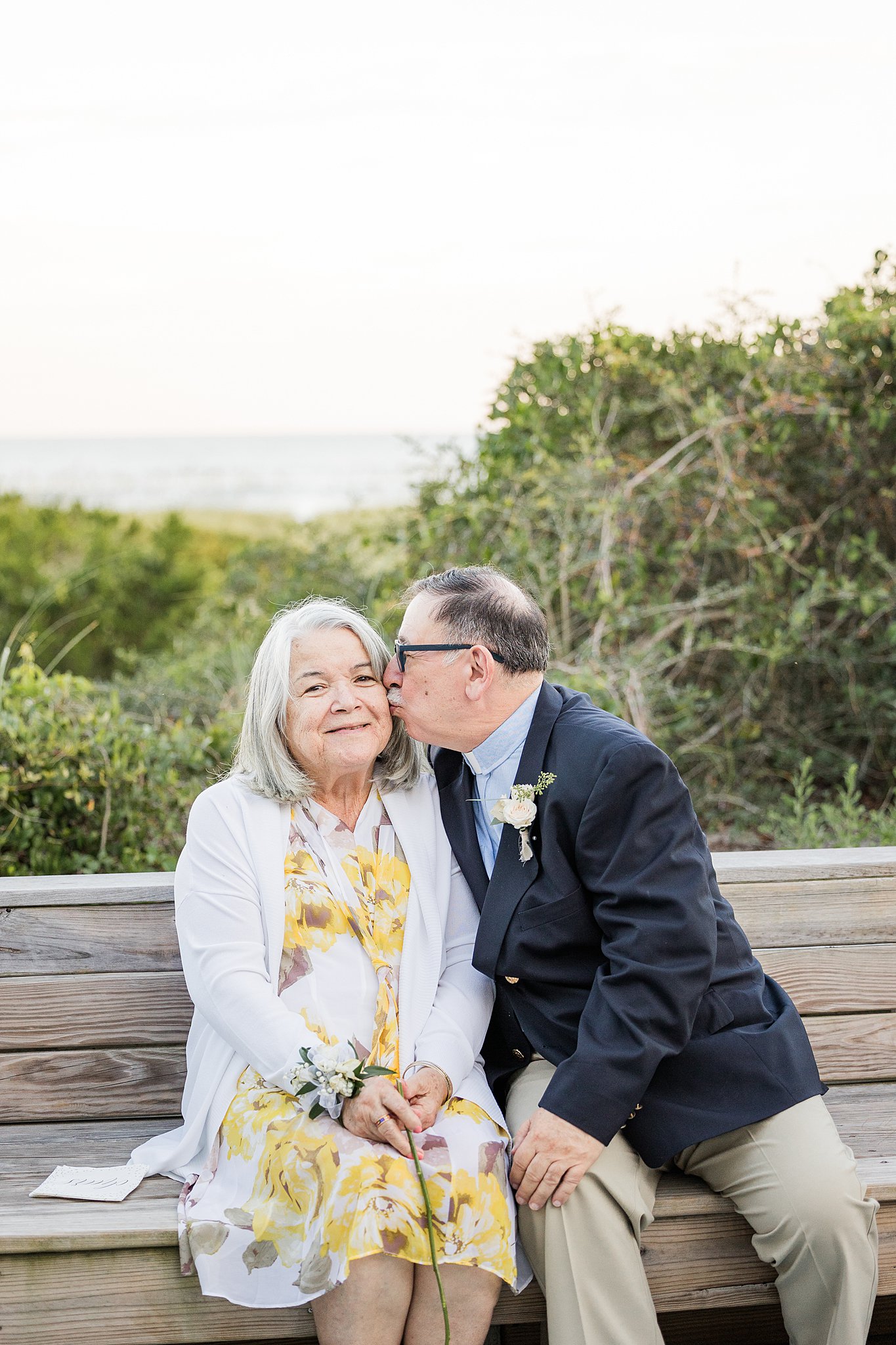 A man sits on a wooden bench by the beach and kisses his wife