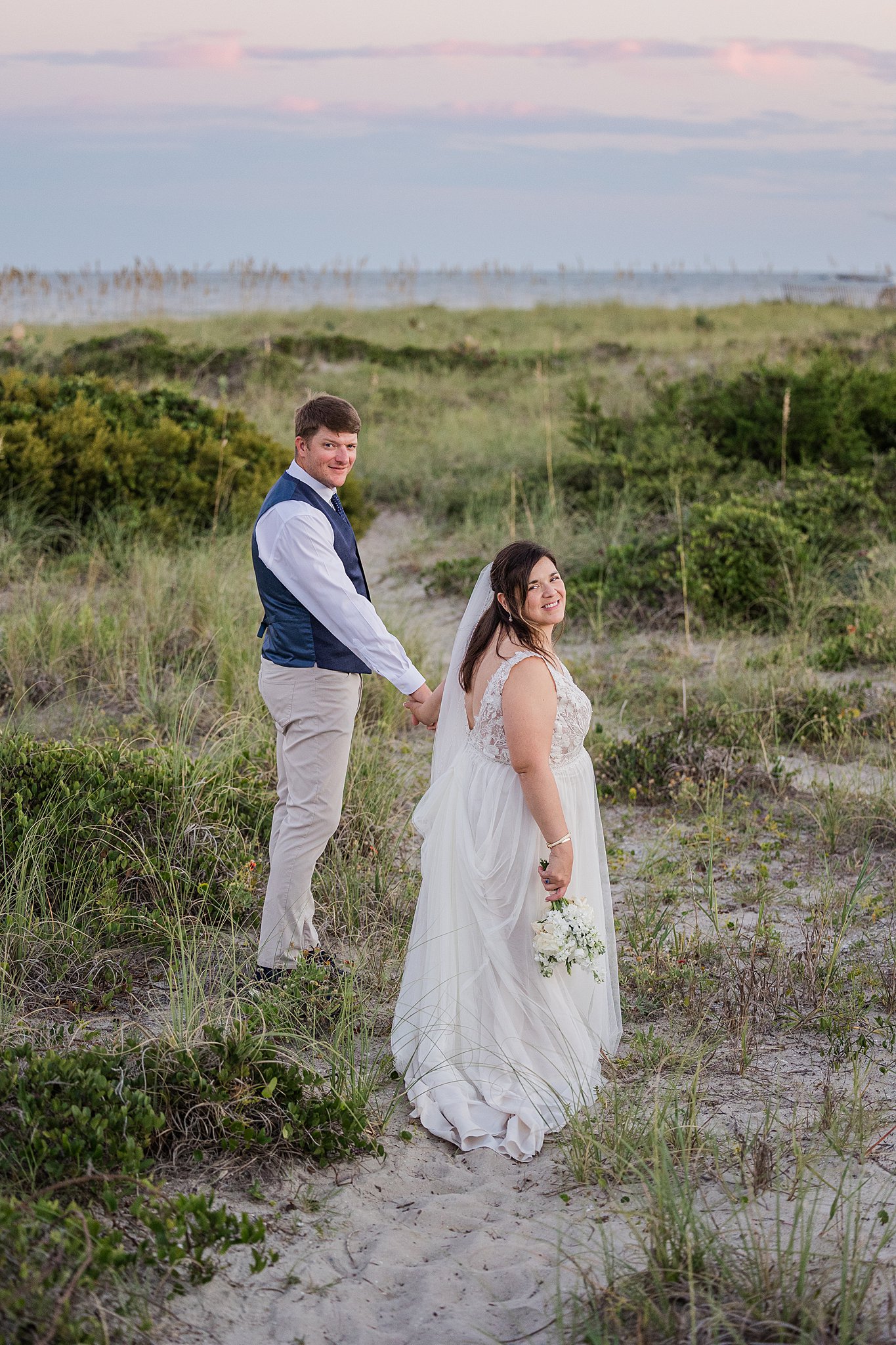 A groom in a blue vest leads his bride down the dune path to the beach at sunset during their wrightsville beach wedding and elopement