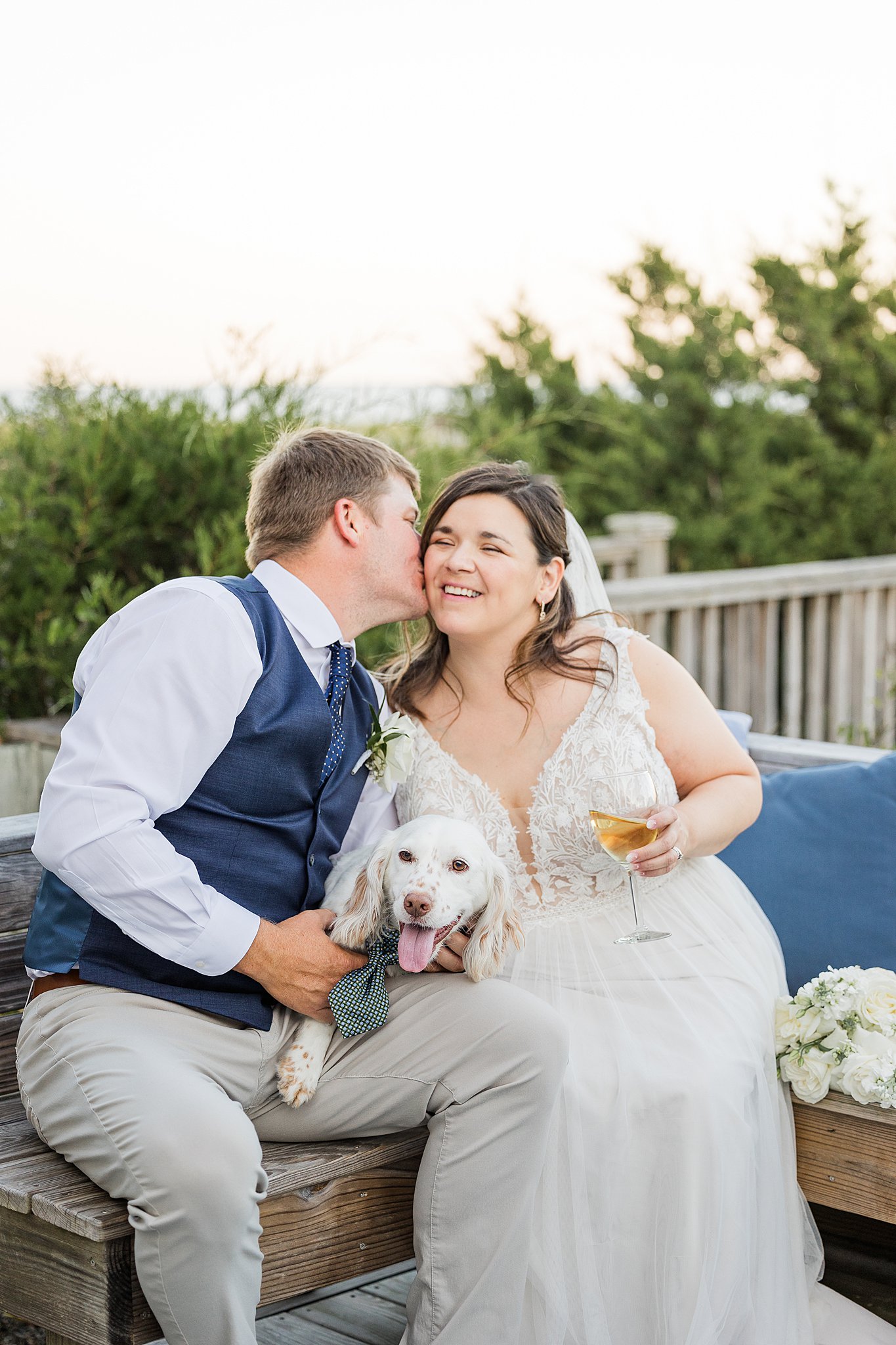 A groom kisses his smiling bride as they sit on a wooden bench with their white dog between them at their wrightsville beach wedding and elopement