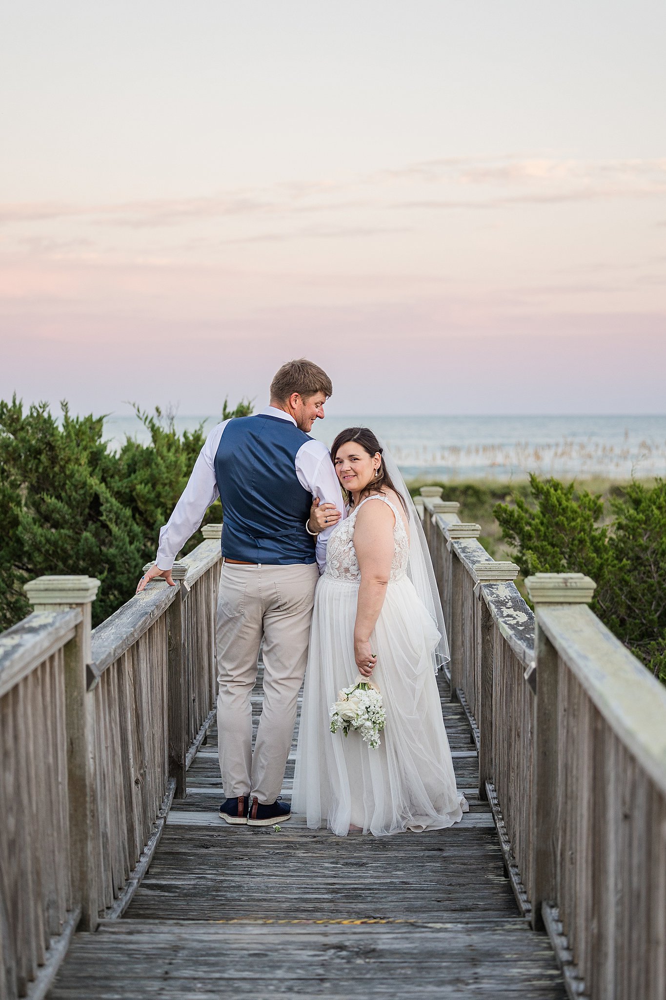 A bride hangs onto the arm of her groom while they walk on a boardwalk during their wrightsville beach wedding and elopement