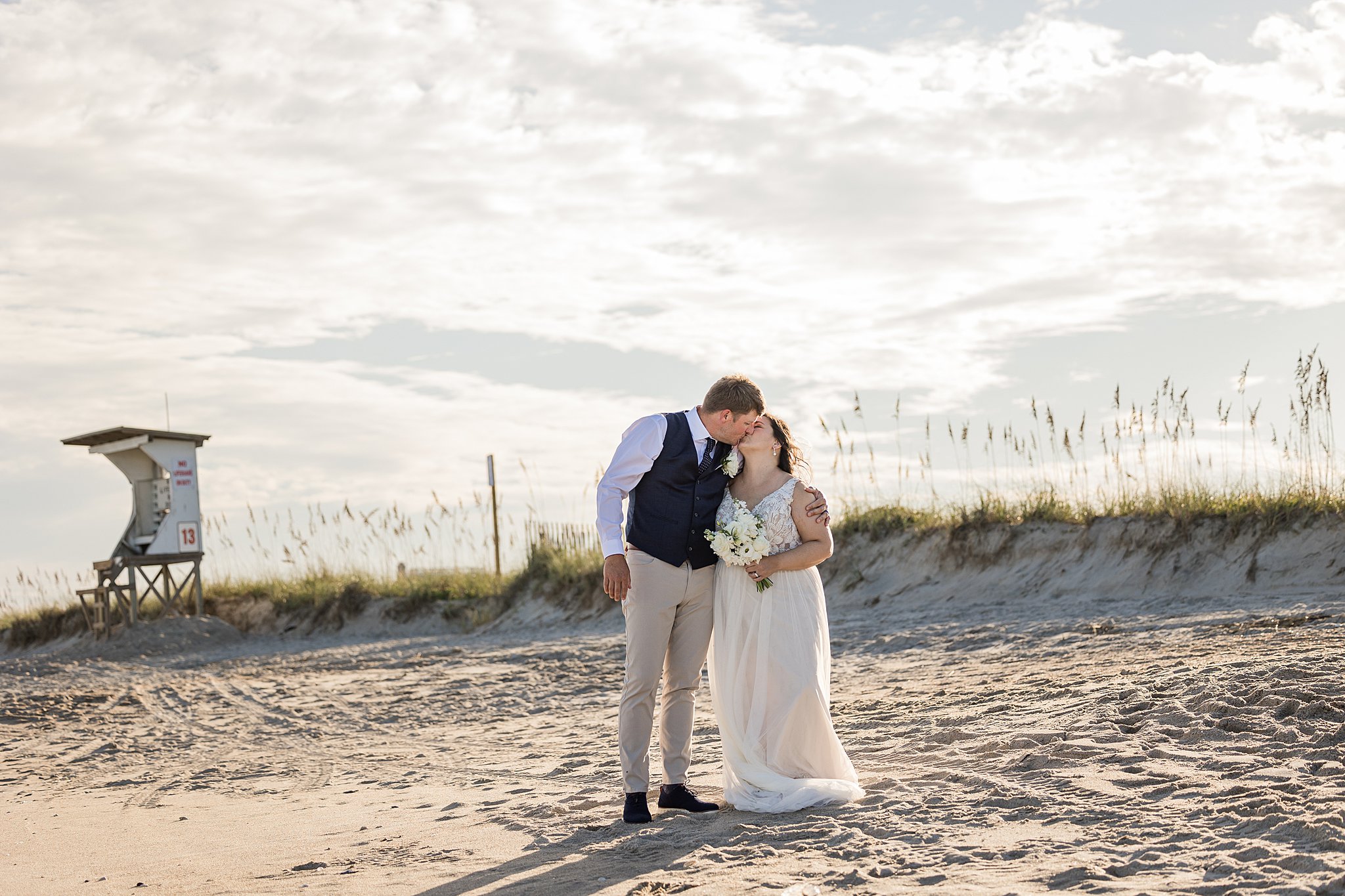 Newlyweds hug and kiss while walking on a beach at sunset during their wrightsville beach wedding and elopement