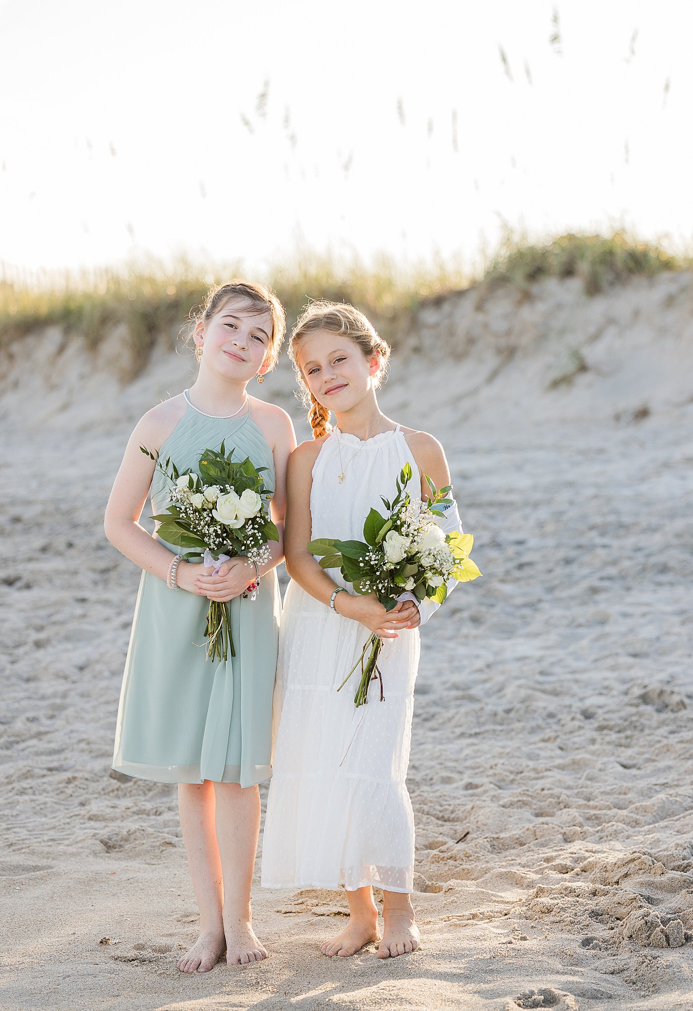 Two young girls in white and green dresses stand on a beach holding white rose bouquets