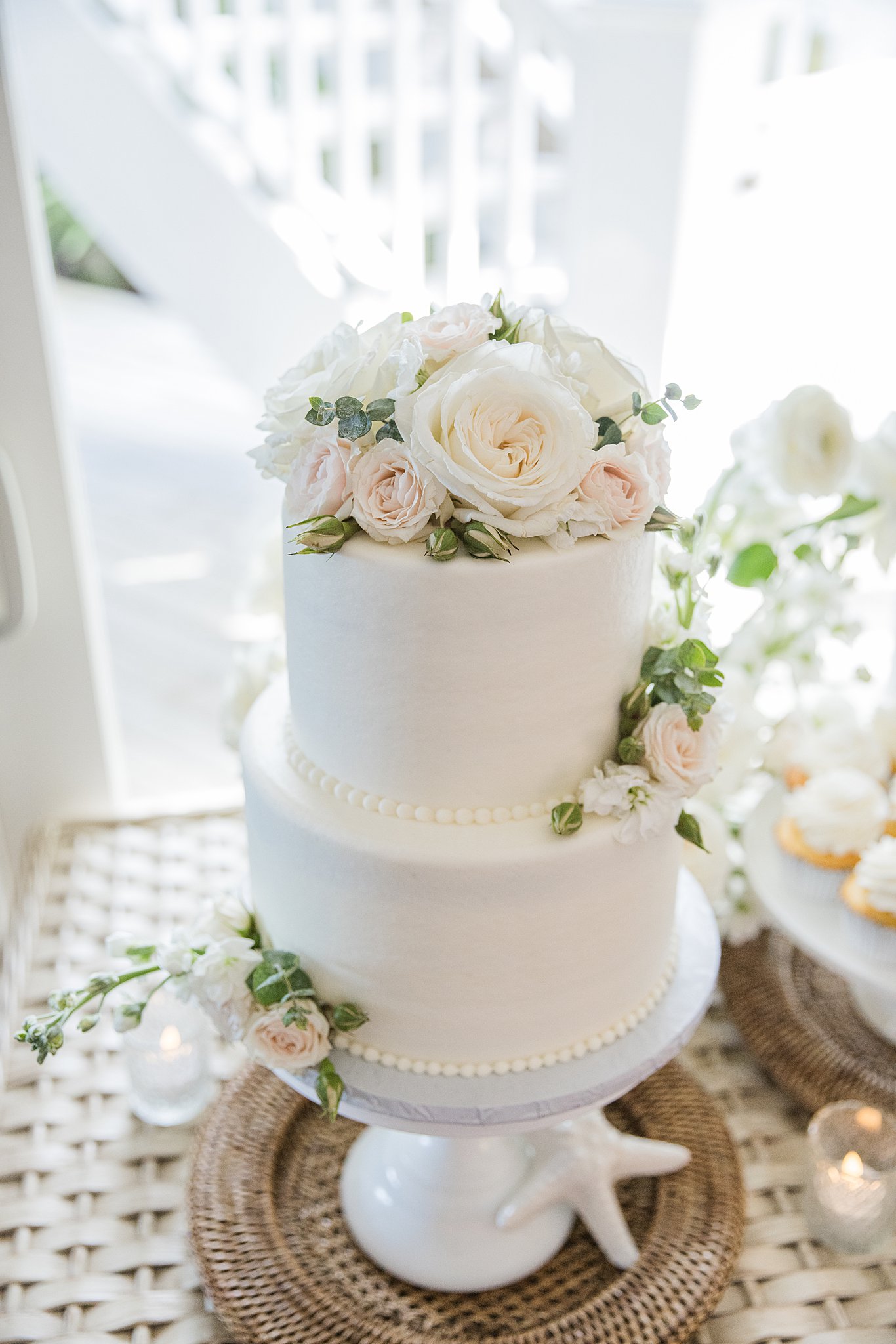 Details of a 2 tier white cake with white roses on a stand and table