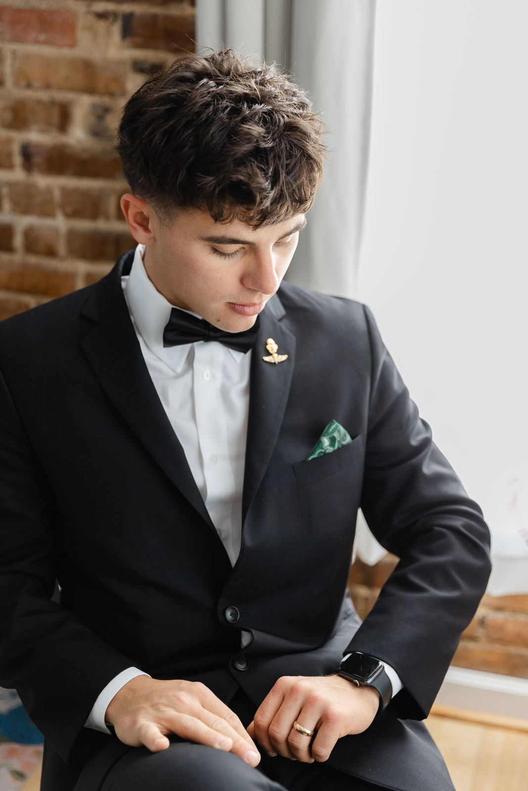 A groom checks his watch while sitting in a window in his black suit