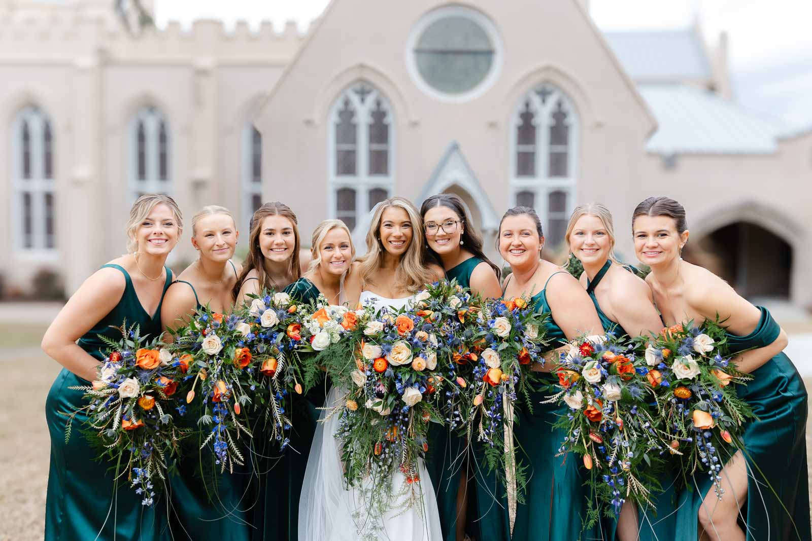 A bride smiles while standing in a lawn with her bridesmaids iin green dresses all holding large colorful bouquets