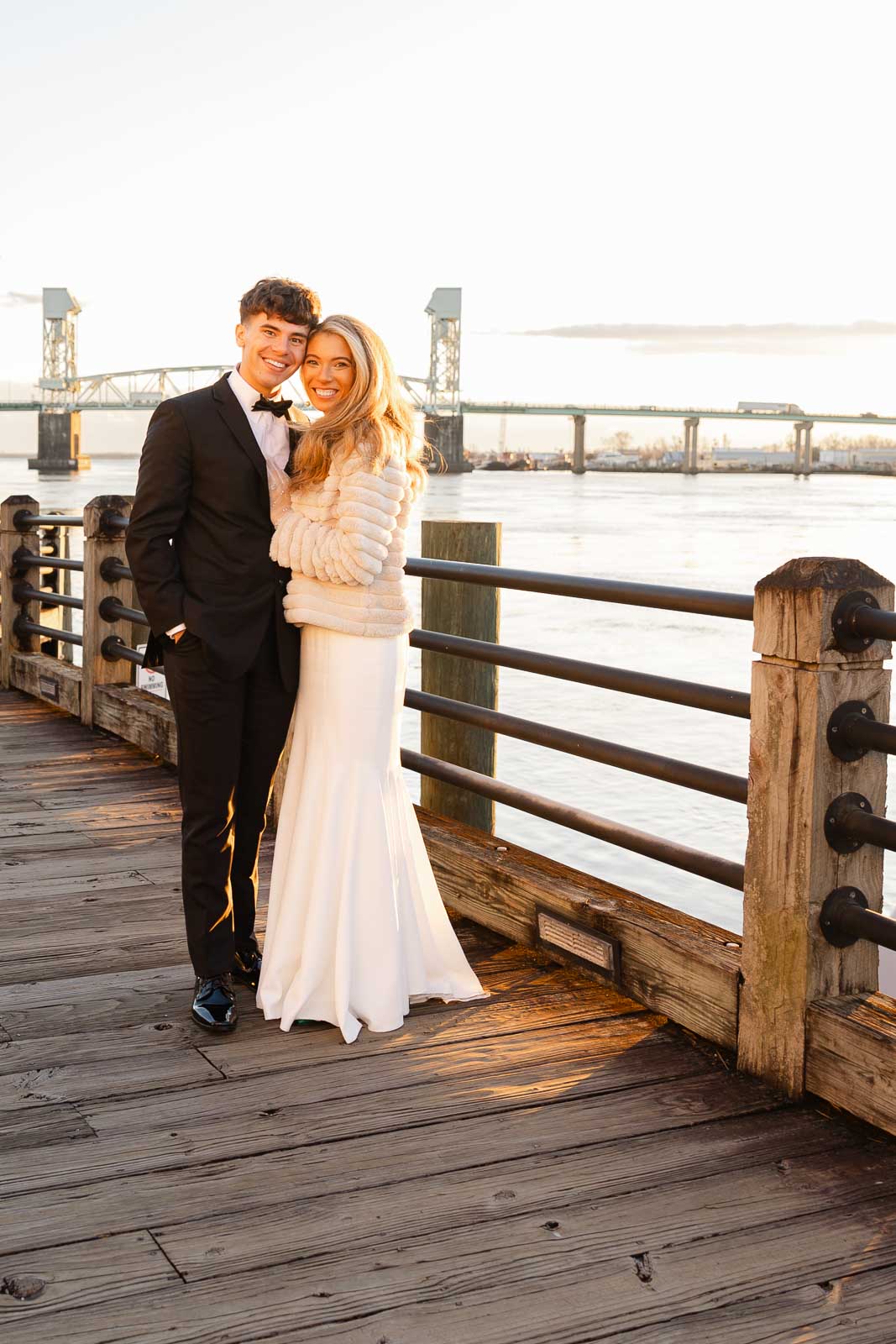 A bride and groom snuggle while walking on a boardwalk at sunset