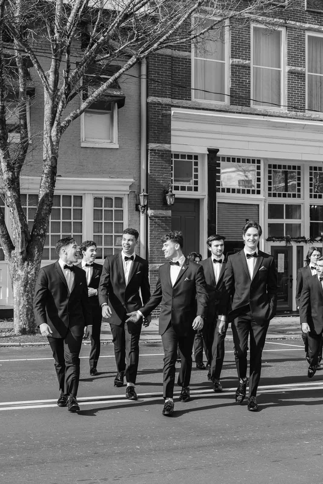 A groom walks across a street while laughing with his groomsmen