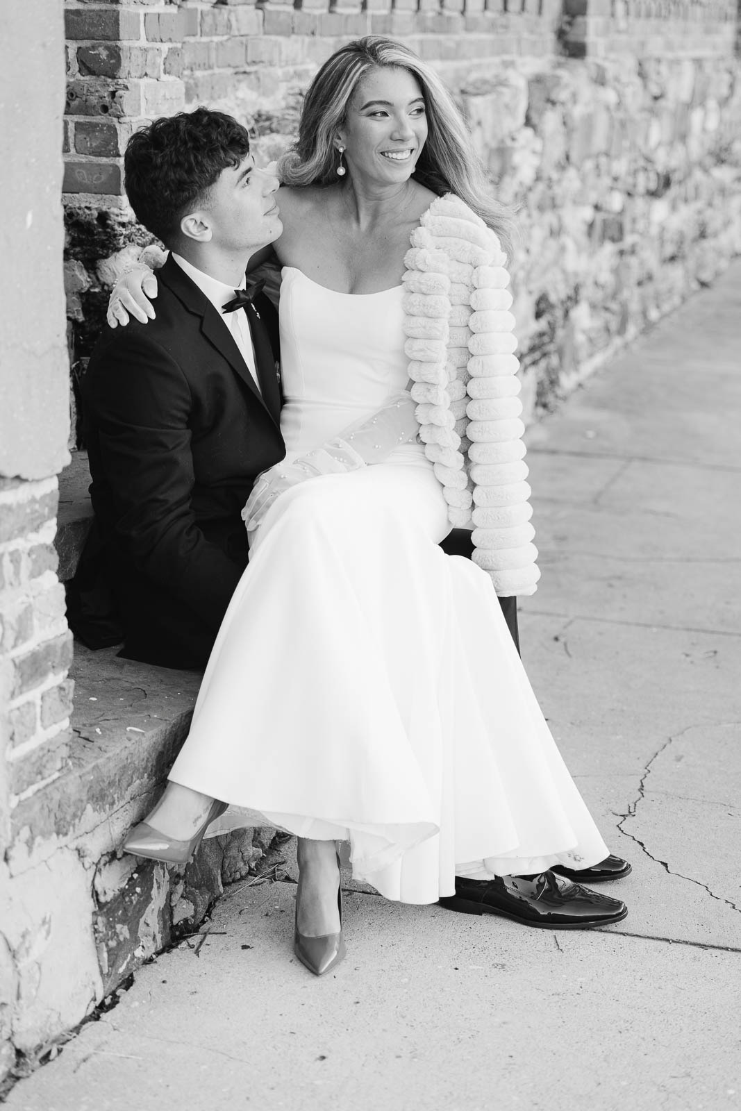 A bride sits in the lap of her groom while smiling over her shoulder on a sidewalk brick step at their Bakery 105 wedding