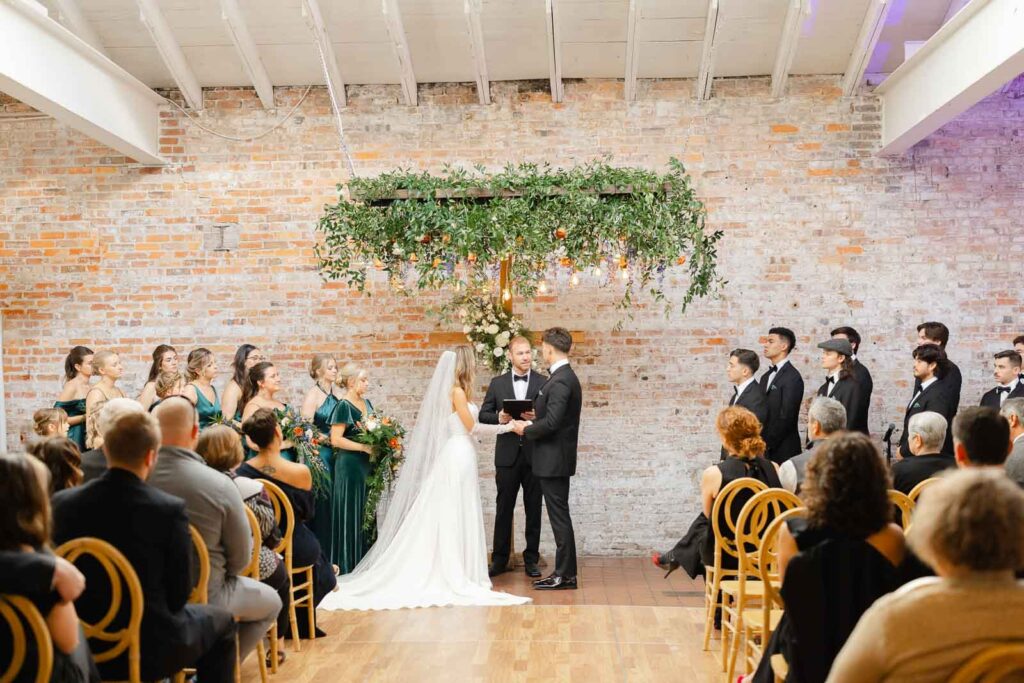 A bride and groom hold hands while listening to their officiant during their Bakery 105 wedding ceremony